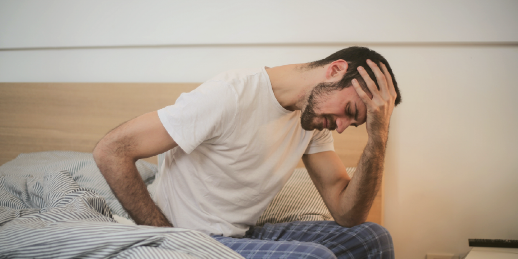 homem jovem sentado na cama , com a mão na cabeça, aparentando sentir dor