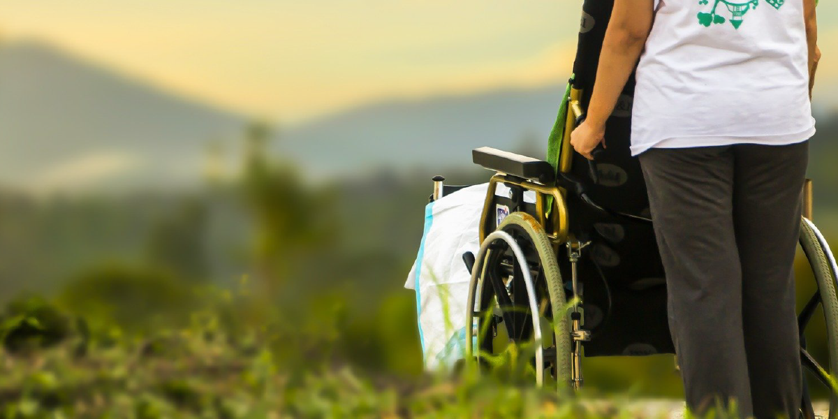 A foto mostra uma mulher levando uma pessoa em uma cadeira de rodas.
