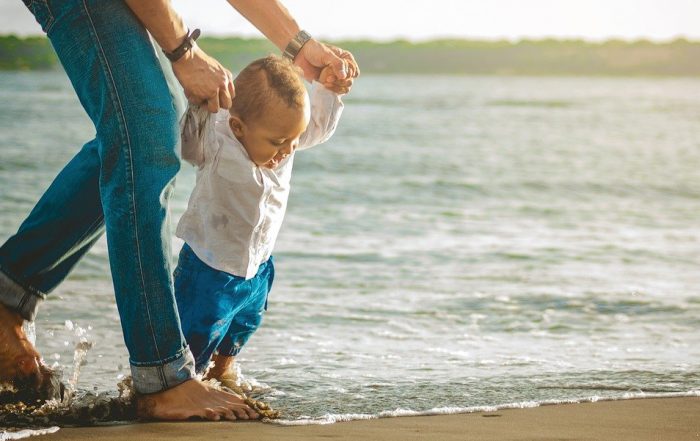 A foto mostra o pai ajudando o filho bebê a caminhar na praia.