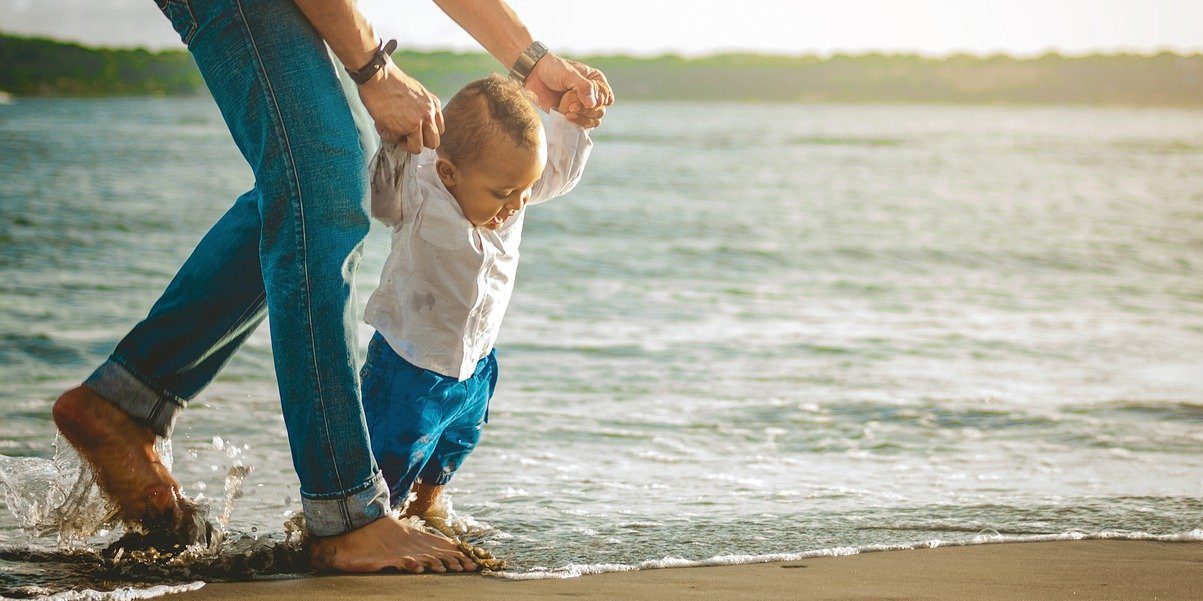 A foto mostra o pai ajudando o filho bebê a caminhar na praia.