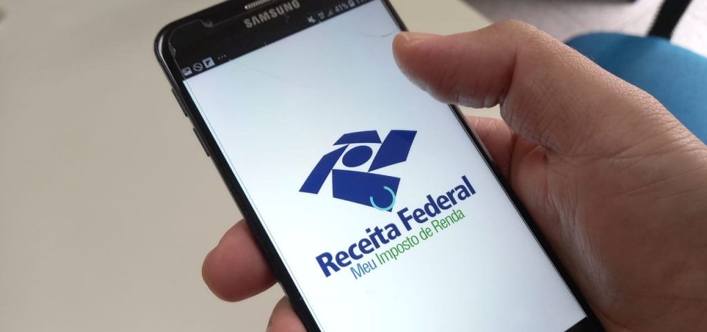 A imagem mostra uma mão segurando um celular com o aplicativo da Receita Federal na tela.