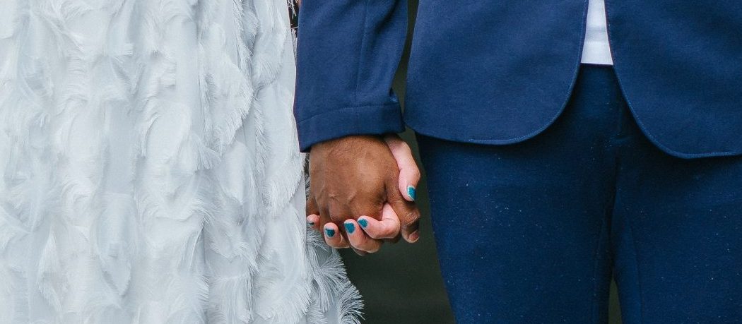 A imagem mostra noivos de mãos dadas, com foco nas mãos deles.