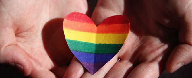 Mãos unidas segurando um coração de papel com as cores do arco-íris, símbolo LGBTQIA+