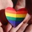 Mãos unidas segurando um coração de papel com as cores do arco-íris, símbolo LGBTQIA+