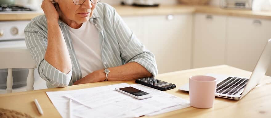 Mulher idosa olhando para uma mesa com papéis, caneta e calculadora.