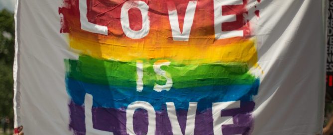 A imagem mostra uma bandeira com as cores LGBTQIA+ dizendo "Love is Love"
