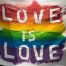 A imagem mostra uma bandeira com as cores LGBTQIA+ dizendo "Love is Love"