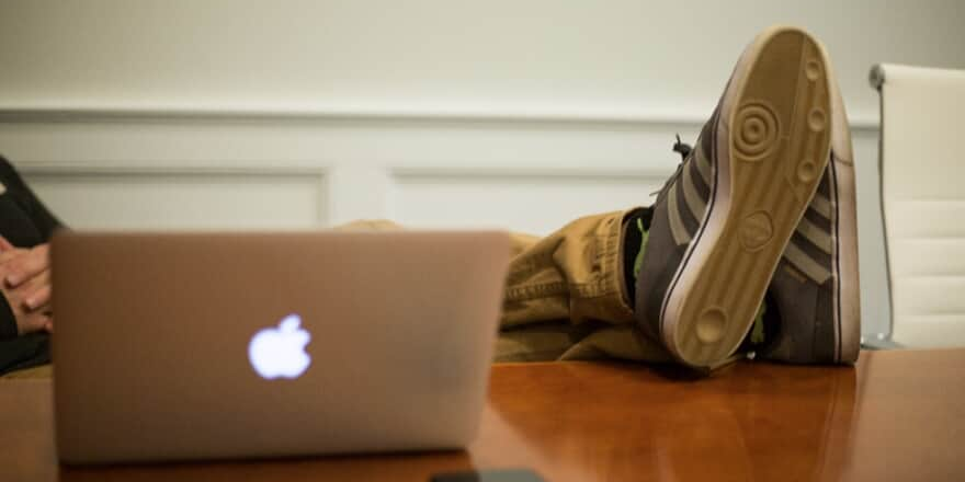 Homem apoia os pés em cima de uma mesa, ao lado de um macbook.