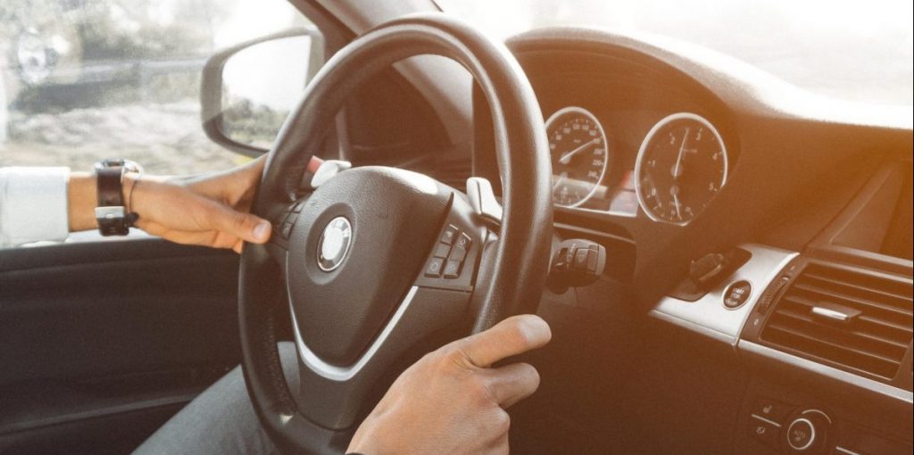 A foto mostra as mãos de um homem ao volante de um carro.