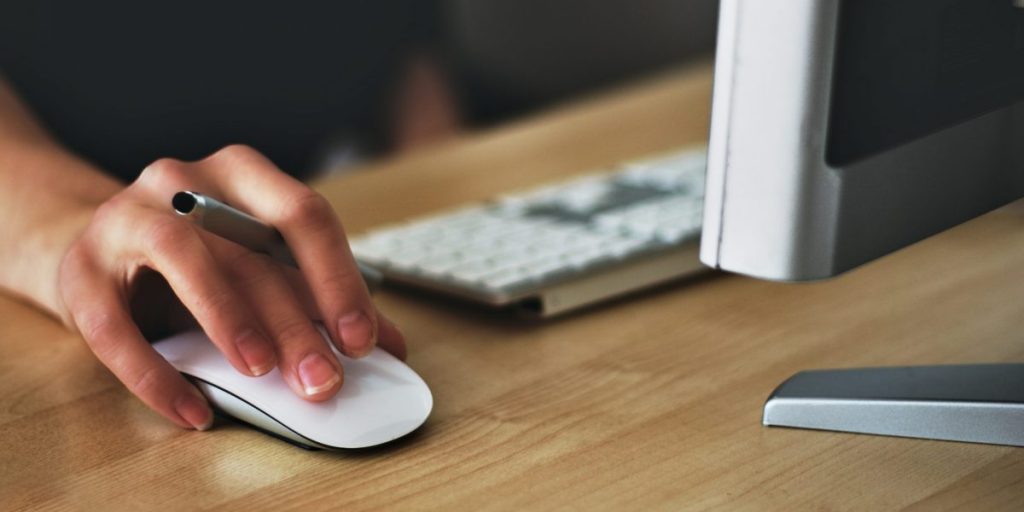 Mão de mulher mexendo em um mouse na frente de um computador.