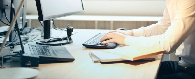 Mão e antebraço de um homem vestindo uma camisa branca apoiados em uma mesa de escritório digitando em um teclado.