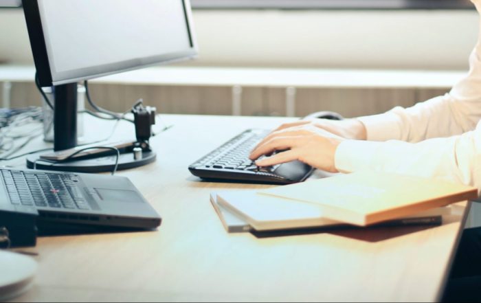 Mão e antebraço de um homem vestindo uma camisa branca apoiados em uma mesa de escritório digitando em um teclado.