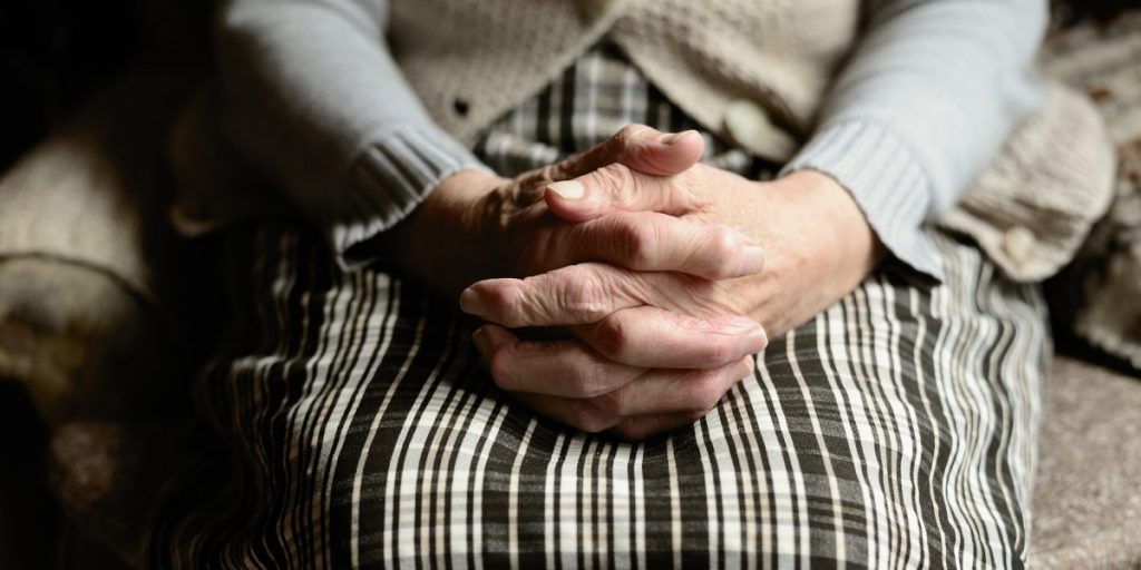 Mulher idosa sentada com as mãos juntas em cima do colo.